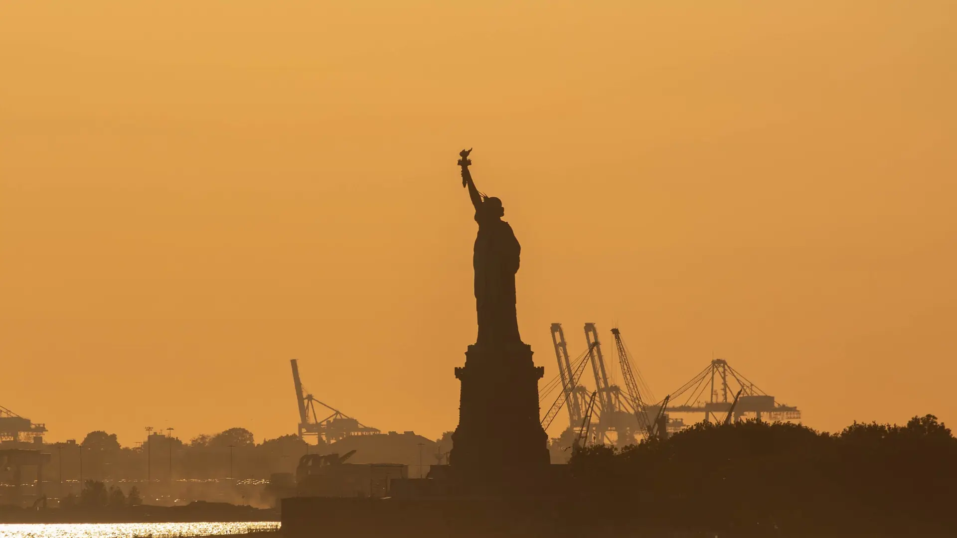 The Statue of Liberty in silhouette at sunset, with harbor cranes on the New Jersey shore behind it